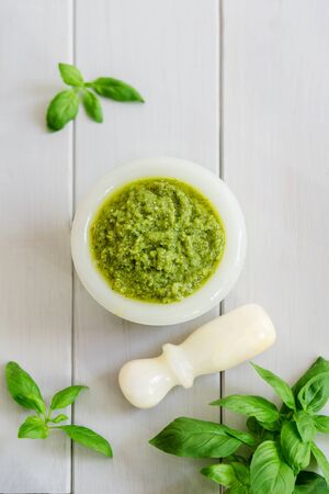 Green italian homemade sauce pesto in stone pounder, made of ground basil, garlic, pine seeds, olives and pecorino sardo cheese. Top view on a white wooden backgroundの写真素材