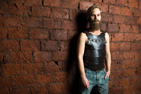 The young man with long hair costs at a red brick wall in a leather vest and jeansの写真素材
