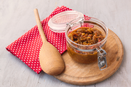 Open jar of chutney of apples, tomatoes, onions and spices on a round cutting board on a gray table.の写真素材