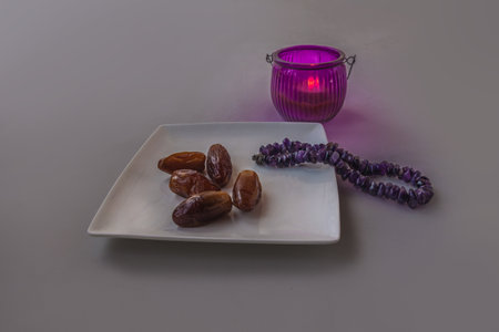 Still life for Ramadan featuring a white plate with dates, amethyst prayer beads, and a glowing purple lantern candle. Simple, spiritual composition symbolizing tradition and fasting. Space for text.の写真素材