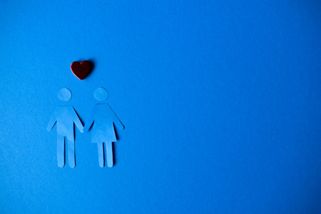 Paper cutouts of a man and woman holding hands with a red heart on a solid blue background.