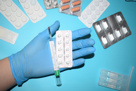Hand in medical gloves holding a thermometer and pills on blue background