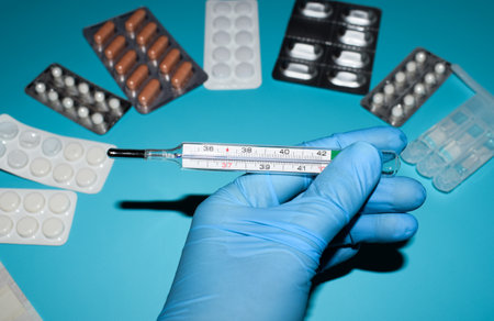 Medical thermometer in hand on a blue background with pills and capsulesの写真素材
