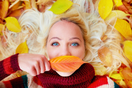 young girl lying among autumn leaves, hiding lips behind a leaf