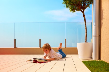 young boy, kid reading book on rooftop terrace, while lying down on deckingの写真素材