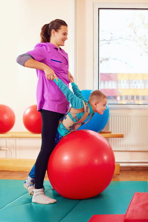 cute kid with disability has musculoskeletal therapy by doing exercises in body fixing belts on fit ballの写真素材