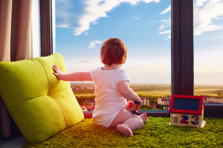 infant baby sitting on carpet at home and watches the beautiful cityscape through the windowの写真素材