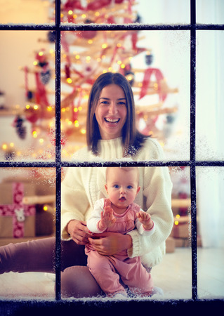 happy mother and baby girl enjoying snowy winter through the window, christmas holidaysの写真素材