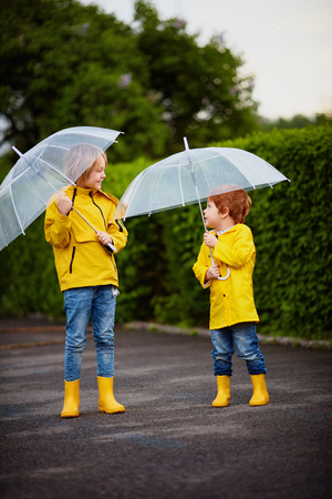 happy young boys, brothers with umbrellas and rain coats and boots walking in spring park at rainy dayの写真素材