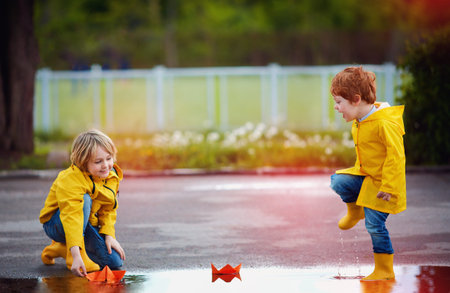 cute boys, brothers having fun together, launching the paper boats in spring puddles, wearing...