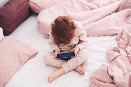 cute redhead baby boy in pajamas playing video games on the phone at the bedroom in the morningの写真素材