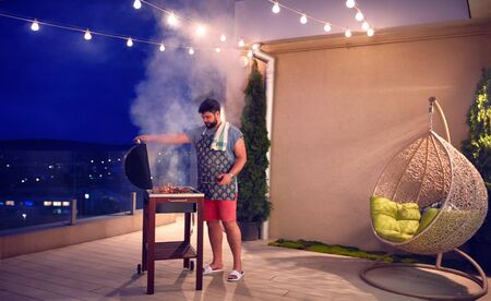 happy adult man grilling the barbecue on rooftop terrace at home in the evening