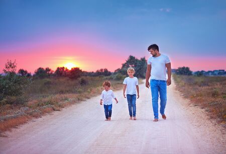 barefoot father and sons walking together along a countryside summer road at sunset. happy familyの写真素材