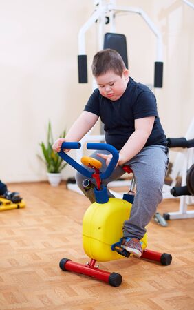 cute kid with down syndrome exercising on stationary bike at the inclusive sport centerの写真素材
