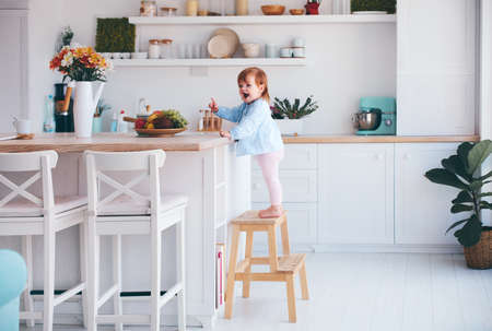 funny infant baby girl standing on a step stool at the cozy kitchen at homeの写真素材
