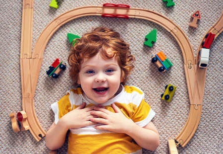 adorable happy redhead baby boy laying on the carpet among railway tracks and trains, wooden toysの写真素材