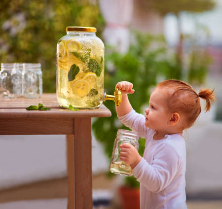 toddler baby girl is trying to pour a fresh citrus lemonade from juice dispenser in the summer gardenの写真素材