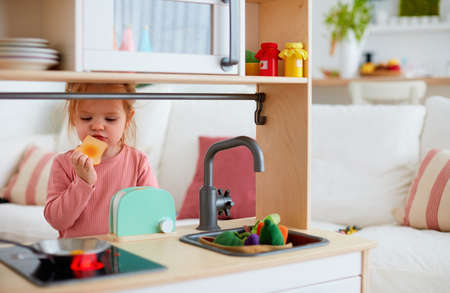 cute toddler baby girl playing on toy kitchen at home, pretends eating a toastの写真素材