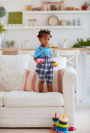 cute african american baby girl playing toys, standing on sofa at the cozy living roomの写真素材