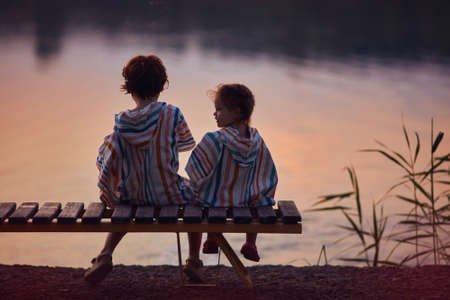 cute children, siblings sitting on the bench by the lake at warm summer eveningの写真素材