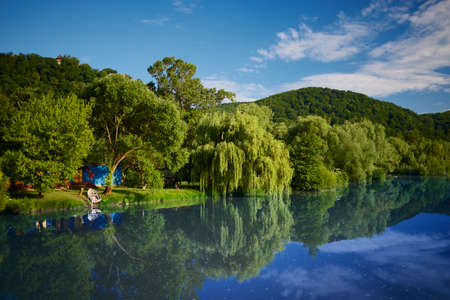 beautiful landscape of blue lake surrounded by lush trees with moored wooden boat and blue houseの写真素材