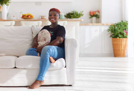 cheerful young woman sitting on sofa in sunlit living room at homeの写真素材