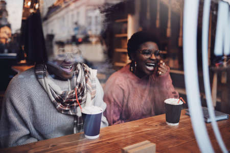 Happy Friends, Laughing African American Women Sitting in Cozy Cafe on Winter Holidaysの写真素材