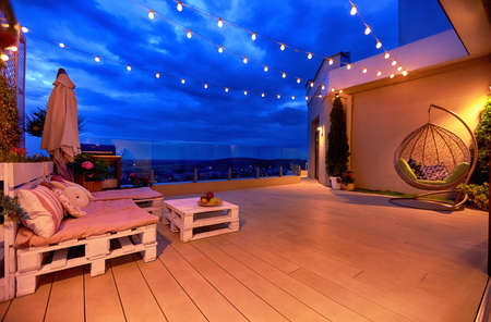 cozy rooftop patio area with lounge zone, hanging chair and and string lights at warm summer eveningの写真素材
