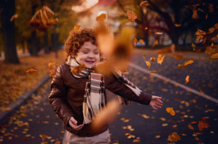 happy boy enjoys autumn swirling around in a flock of falling golden leaves, cozy city street. Focus on leavesの写真素材
