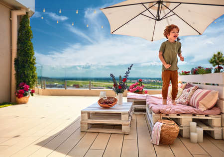 smiling kid having fun, jumping on lounge area made of wooden pallets, summer rooftop patioの写真素材