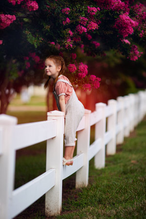 happy redhead baby girl on the fence enjoys the blooming branches of crape myrtle trees. Beautiful lagerstroemia in bloomの写真素材