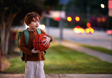 portrait of cute redhead school boy, kid with backpack and the ball on the evening streetの写真素材
