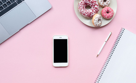 Smartphone with empty screen on pink office desk background, surrounded with laptop, plate with donuts and notebook.の写真素材