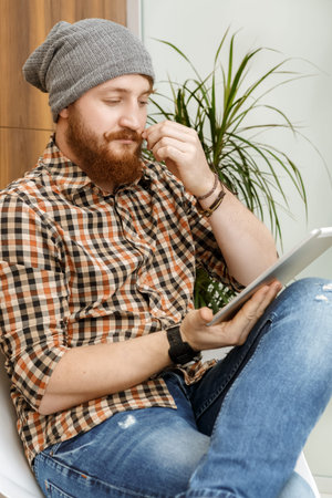 Handsome red-haired hipster thoughtfully reading a book on  tablet