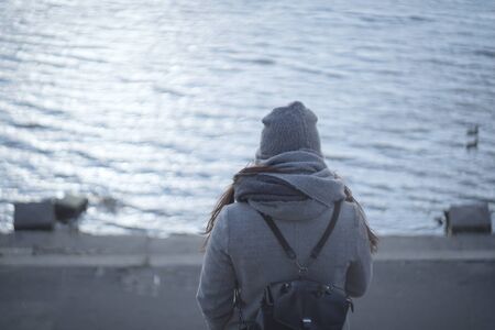 Back view of brunette woman in grey coat standing on river bank or lake shore. Caucasian girl enjoying autumn day outdoors. Nature, water, lifestyle.の写真素材