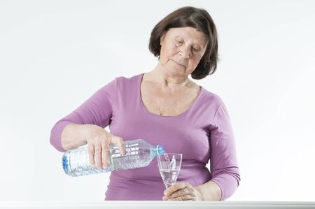 Elderly woman pours water into a glass.Studio photography on a light gray background.の写真素材
