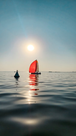 A sailboat with a vibrant red spinnaker sail glides smoothly across calm water under a clear sky. Silhouettes of people are visible on the boat, enjoying a peaceful sailing experience. The sun reflects off the water, creating sparkling highlights and a serene atmosphereの写真素材