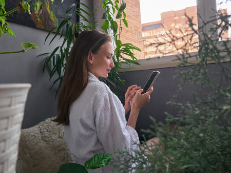 Woman in Robe Checking Phone in Cozy Balcony Gardenの写真素材