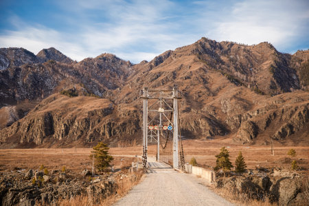 Old suspension bridge on a dirt road leading towards vast mountains. Rural landscape adventure travel concept for nature exploration.の写真素材
