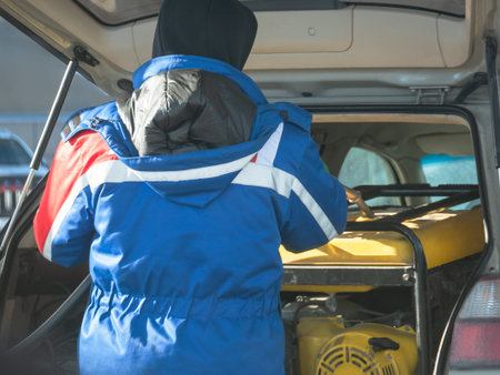 Man in blue jacket putting a yellow industrial generator into a car trunk. Emergency power equipment...