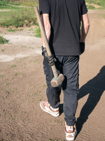 Man walking away on a dirt road while carrying a heavy sledgehammer in his hand. Concept of...