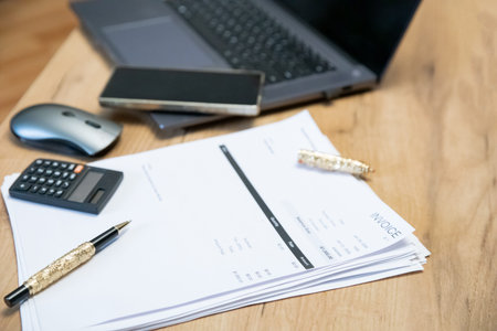 Stack of invoice documents with a pen, calculator, and laptop on a wooden desk. Business financial...