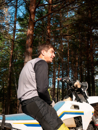 Caucasian man motocross racer sitting on motorcycle in forest. Rider resting during extreme trail...
