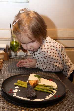 A little girl in a restaurant looks at what the official brought on a plate with baked salmon and asparagus, decorated with sauce and caviar of flying fish, on the table in front of her she looks very carefully at how the fish dish is decorated Her daughter looks at the food with hungry eyes and wants to try it as quickly as possibleの写真素材