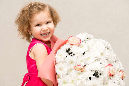 A little girl in a bright pink dress with curly hair is holding a huge bouquet of chrysanthemums and roses for her mother and smiling broadly and fervently.の写真素材