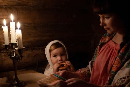 in a Russian hut of tsarist Russia, a young woman is doing needlework at a table and a child is sitting next to her and eating a bagel. An old candelabra with candles is burning on the tableの写真素材