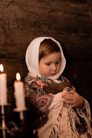 Russian hut of tsarist Russia, a little girl in Russian national clothes leans against a log wall and hugs an ancient book of Holy Scripture. The kid is thinking about something, an old candelabra with candles is burning on the table. The girl thinks about what she has readの写真素材