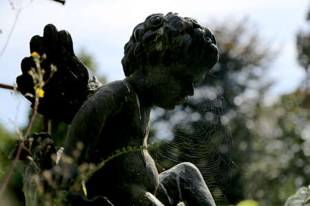 Beautiful Cupid angel statue with wings on back in vintage garden. Cupid angel statue consisting of old dark stone. Interior of abandoned garden is big fine Cupid angel statue, green leaves on trees.の写真素材