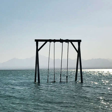Wooden childrens swing stands in blue calm sea under open clean sky. Swing consists of seat hanging on thick ropes for jumping into sea. Baby swings flooded sea for people on background mountains.の写真素材