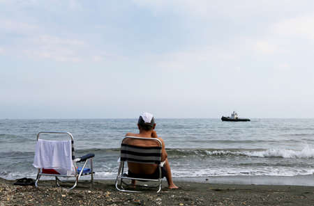Man sitting on chair is watching an old ship sailing on sea. Old ship floating in water sees person resting on chair. To chair can see male as which is watching old ship sails along horizon of nature.の写真素材